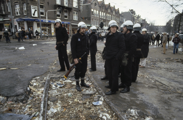 Eine Gruppe von Polizisten in Helmen und mit Waffen in der Hand steht an der Seite einer Straße. Im Hintergrund sind Gebäude mit Fenstern, Schilder mit Text, Laternenpfähle, Bäume und ein klarer Himmel zu sehen.