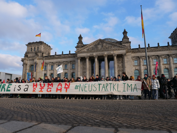 Gruppe von Menschen vor dem Reichstag in Berlin mit einer Fahne, auf der "Wir sind ein Menschenrecht" steht, mit den architektonischen Details des Gebäudes und Flaggen im Hintergrund.