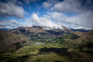 Ein atemberaubender Blick von der Spitze eines Berges in Queenstown, Neuseeland, der saftig grünes Gras, Bäume, eine gewundene Straße und einen Himmel voller weißer, flauschiger Wolken zeigt.