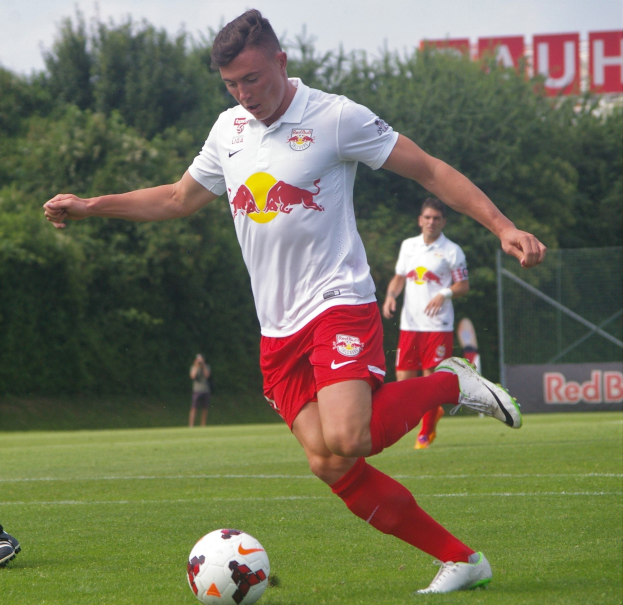 Ein professioneller Fussballspieler in einem RB Leipzig-Trikot schießt einen Fussball auf einem grünen Feld mit Bäumen und einem klaren blauen Himmel im Hintergrund.