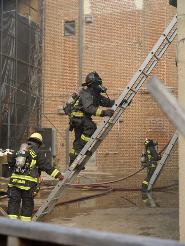 Feuerwehrleute in Helmen und Ausrüstung, die eine Leiter vor einem Backsteinbau hochklettern, mit Rohren auf dem Boden und einer Metallstange am Boden, mit einem weiteren Gebäude und einem Netz im Hintergrund.