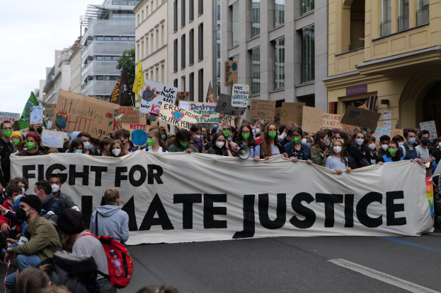 Eine große Gruppe von Menschen marschiert mit einem Banner, auf dem 'Kampf für Hassjustiz' steht, durch die Straße. Einige tragen Masken und halten Schilder, während andere Taschen tragen; im Hintergrund sind Gebäude, Bäume und ein klarer blauer Himmel zu sehen.