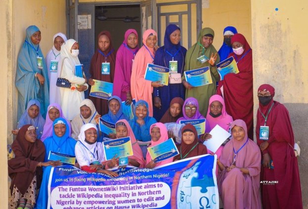 Gruppe von Frauen, die strahlend in die Kamera lächeln und Urkunden in den Händen halten, vor einem Gebäude mit einem Banner, auf dem steht: "Funtua Women Wiki Initiative setzt sich für die Bekämpfung der Wikipedia-Ungleichheit im nördlichen Nigeria ein."