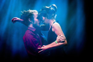 Ein Mann und eine Frau umarmen und küssen sich auf einer Bühne, während ein paar Scheinwerfer den Hintergrund beleuchten.