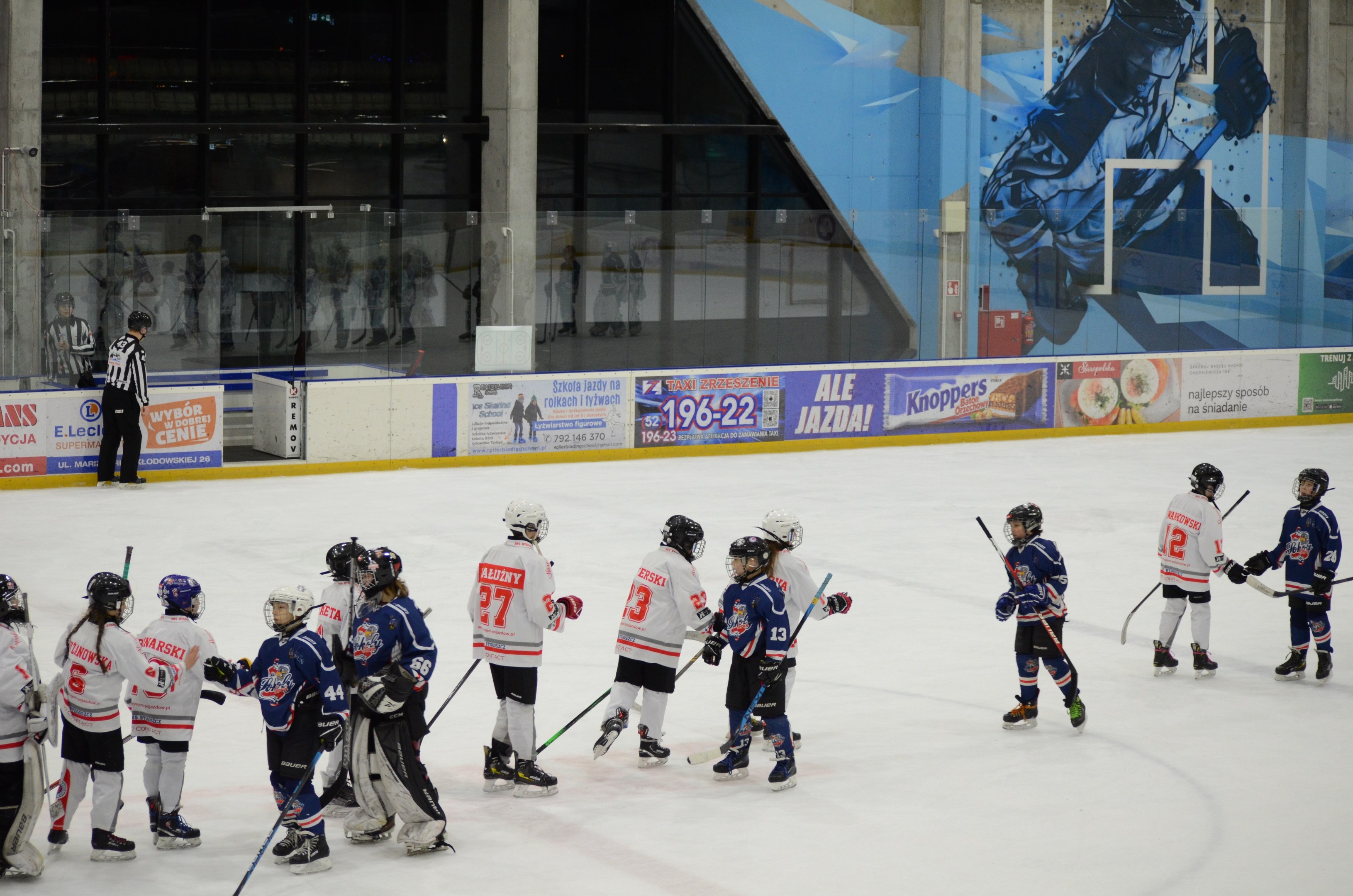 Gruppe junger Menschen, die Eisockey auf einer Indoor-Eisfläche spielen, mit Helmen und Stöcken, einer Person links daneben und Bannern, Säulen und Glaswänden im Hintergrund.