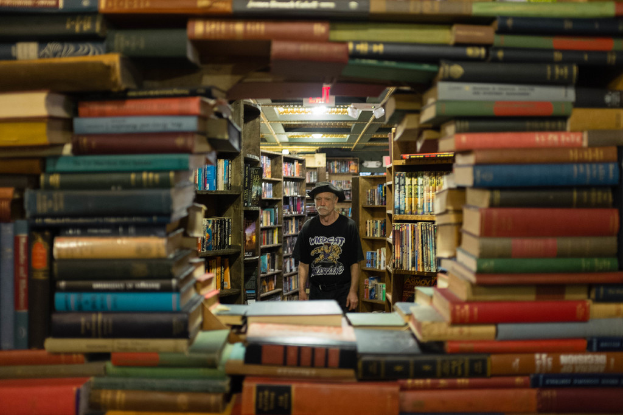 Ein Mann in einem Hut, T-Shirt und Hose steht in einer Bibliothek mit ordentlich aufgestellten Büchern auf Regalen.