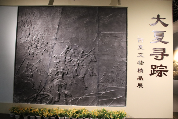 Ein großes steinernes Schild mit chinesischer Schrift darauf, umgeben von Blumentöpfen mit Pflanzen, beleuchtet von Deckenlampen.