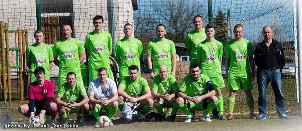 Gruppe von Männern in grünen T-Shirts, die auf einem Fußballfeld stehen oder knien, mit einem Ball, einem Torpfosten auf der rechten Seite und verschiedenen Hintergrundelementen wie Bäume, Gebäude und Fahrzeuge.