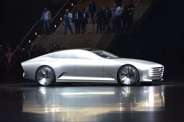 Ein Mercedes-Benz F 015 Luxury in Motion Concept-Wagen, glänzend schwarz mit Chromakzenten, auf einem Boden mit einer Gruppe von formell gekleideten Menschen im Hintergrund auf einer Treppe stehend.