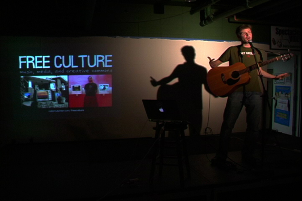 Eine Person, die eine Gitarre hält und vor einem Tisch mit einem Laptop und einer Projektionsfläche mit der Aufschrift "Free Culture" steht.