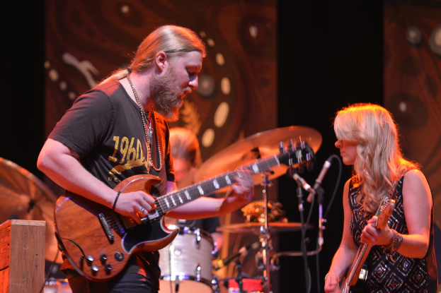 Ein Mann und eine Frau spielen Gitarren, im Hintergrund sind Trommeln zu sehen.