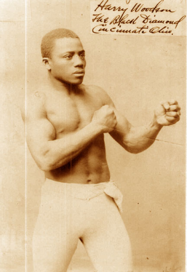 Ein Mann in einem Boxring mit Handschuhen und Shorts, der entschlossen aussieht und aufrecht steht, mit Text auf dem alten Foto.