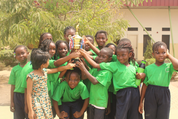 Kinder stehen zusammen und halten einen Pokal in einer Schulhofumgebung mit Bäumen, Pflanzen und einem Gebäude im Hintergrund.