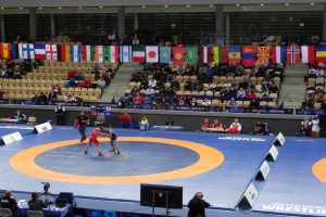 Zwei Wrestler im Ringkampf in einem vollen Stadion mit Zuschauern, Fotografen und Equipment im Hintergrund.