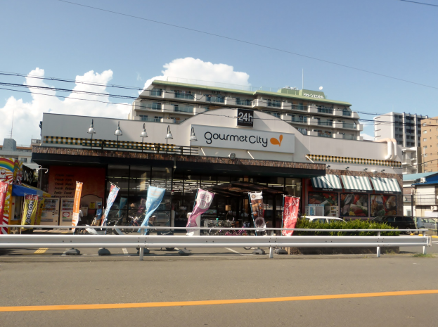 Eine belebte Stadtstraße mit Gebäuden, Straßenlaternen, Fahrzeugen und einem Restaurant mit Essensauslage im Schaufenster.