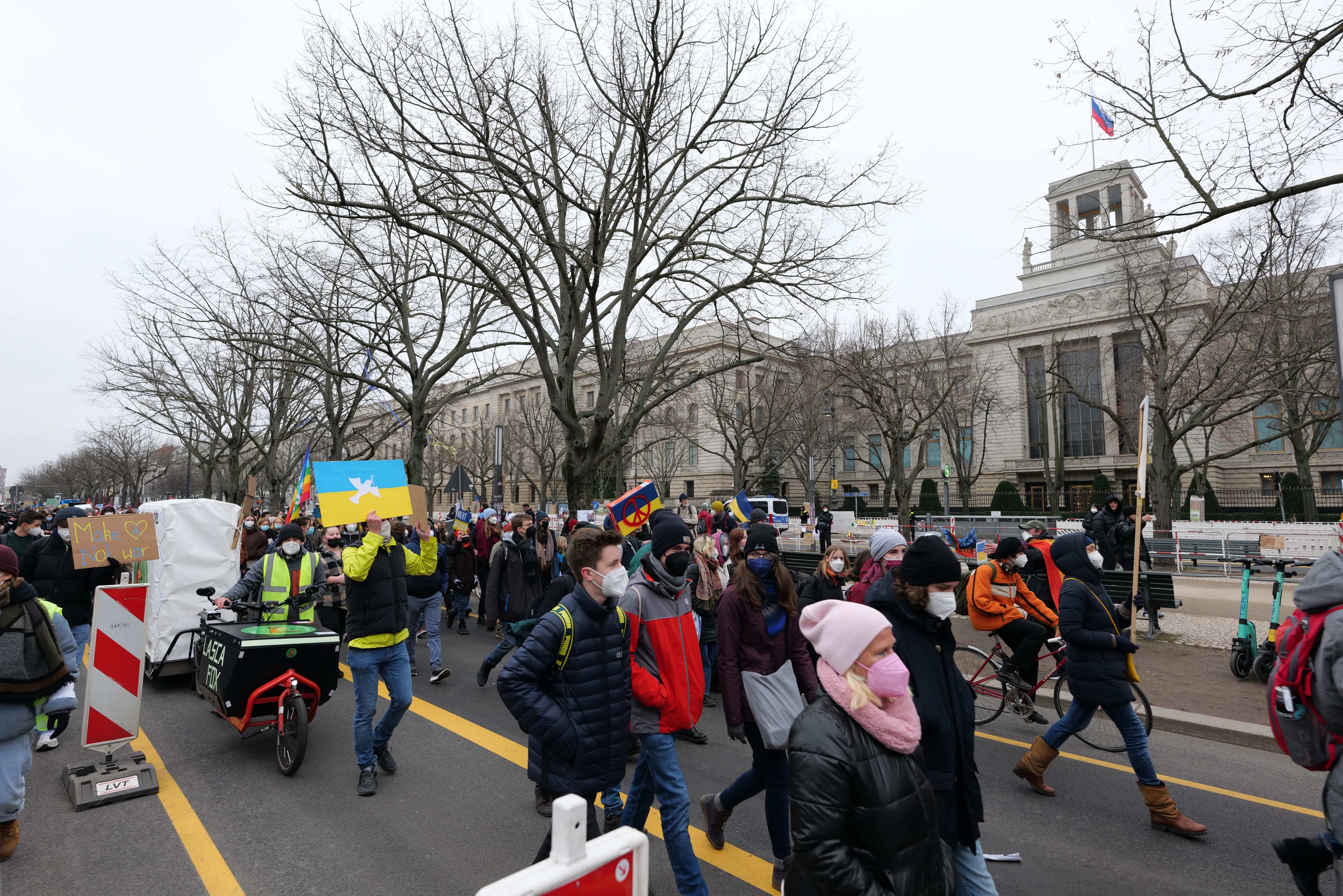 Eine große Gruppe von Menschen nimmt an einer Protestdemo auf einer Straße in Washington, D.C. teil, einige halten Schilder und Banner, andere fahren Fahrräder, mit Bäumen, Schildern und einem klaren blauen Himmel im Hintergrund.