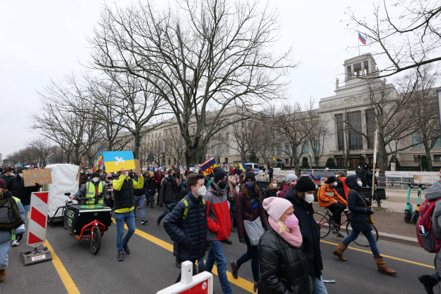 Eine große Gruppe von Menschen nimmt an einer Protestdemo auf einer Straße in Washington, D.C. teil, einige halten Schilder und Banner, andere fahren Fahrräder, mit Bäumen, Schildern und einem klaren blauen Himmel im Hintergrund.