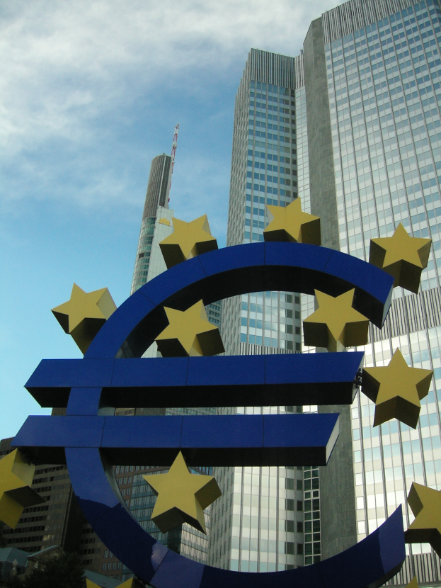Das Euro-Zeichen vor der Europäischen Zentralbank in Frankfurt, Deutschland, umgeben von Bäumen unter einem bewölkten Himmel.