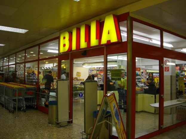 Ein Supermarkt mit einem Eingang in der Mitte, Einkaufswagen auf der linken Seite und einigen Einkäufern, die Produkte betrachten.