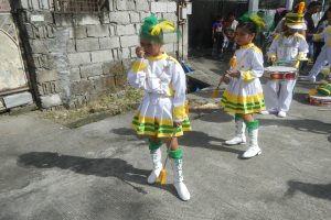 Eine Gruppe junger Mädchen in weißen und gelben Outfits marschiert, während sie Musikinstrumente spielen, umgeben von einer Wand, einem Tor und Bäumen.