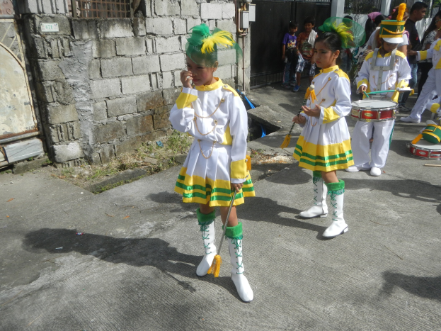 Eine Gruppe junger Mädchen in weißen und gelben Outfits marschiert, während sie Musikinstrumente spielen, umgeben von einer Wand, einem Tor und Bäumen.