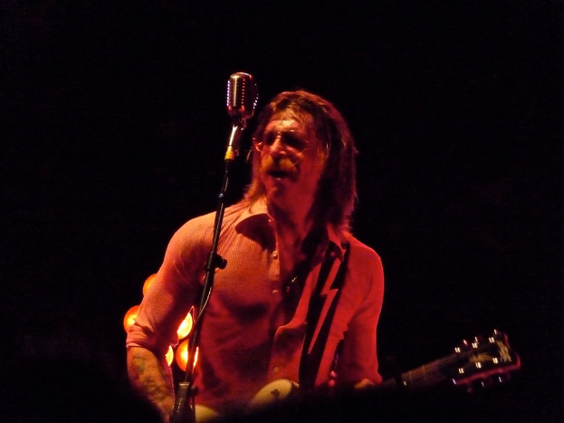 Ein Mann mit Brille steht und singt, während er eine Gitarre hält und seinen Mund öffnet.