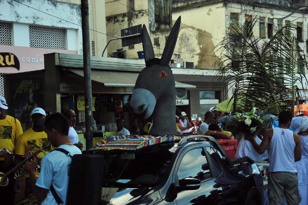 Eine Skulptur in der Mitte mit Menschen drumherum, Gebäuden und Drähten im Hintergrund, einem Blumenstrauß, einem Auto, einem Pfahl, einer Person, die auf der linken Seite ein Musikinstrument spielt, und einem Geschäft.