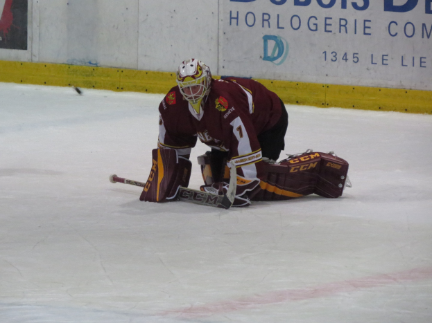 Eishockeyspieler in rot-gelber Uniform, der einen Schuss auf dem Eis hält, trägt Helm, Handschuhe und Knieprotektoren und hält einen Eishockeyschläger, mit einer Wand und Text im Hintergrund.