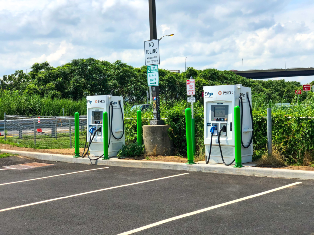 Elektrische Fahrzeug-Ladestation auf einem Parkplatz mit umgebenen Infrastruktur, Grünflächen und einer Brücke im Hintergrund.