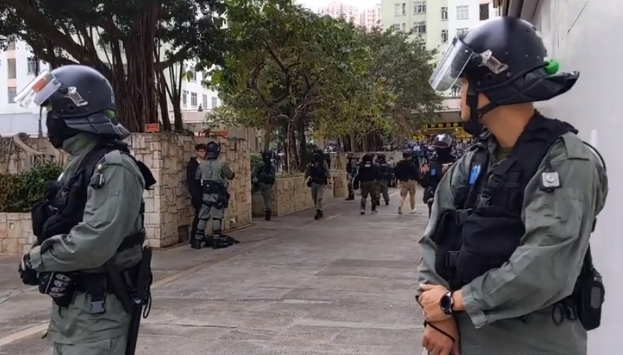 Eine Gruppe von Polizisten in Helmen und Schutzwesten steht auf einer Straße in Hong Kong, mit Bäumen, Gebäuden und Himmel im Hintergrund, während sie versuchen, Demonstranten zu zerstreuen.