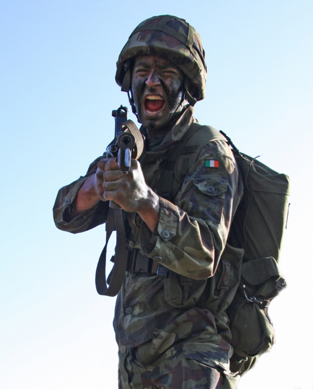 Eine Person in militärischer Uniform, mit einem Abzeichen, Helm und einer Tasche, hält eine Waffe.