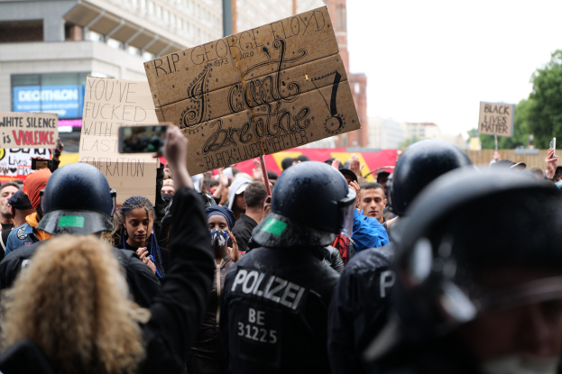 Gruppe von Menschen mit Helmen und Schildern bei einer Demonstration, mit Gebäuden, Bäumen und einem Mast im Hintergrund.