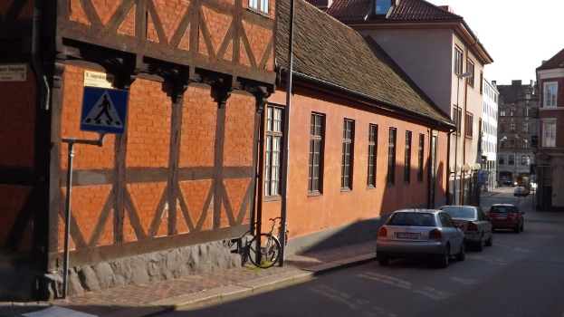 Fahrzeuge auf der Straße mit Gebäuden, Laternen, Verkehrsschildern, Fenstern und Dächern im Hintergrund und einem Fahrrad auf dem Gehweg neben einem Pfahl.
