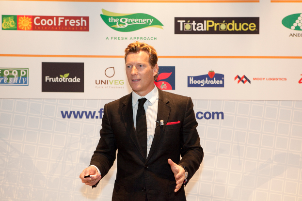 Ein Mann in einem schwarzen Blazer steht und hält ein Objekt, während er spricht, mit einem weißen Banner und einer weißen Tafel mit organizationalen Logos im Hintergrund.