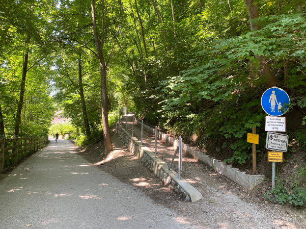 Eine Person fährt mit dem Fahrrad auf einem asphaltierten Weg durch den Wald, umgeben von Bäumen und einem Holzzaun auf der linken Seite und Schildern mit Text und Bildern auf der rechten Seite.