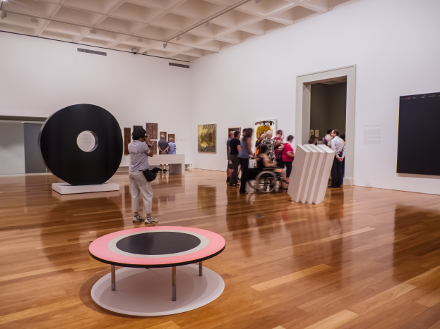 Eine Gruppe von Menschen in einer Kunstgalerie mit Fotorahmen an den Wänden und Deckenlampen, mit einem Menschen im Rollstuhl in der Mitte und einem schwarzen Objekt auf der linken Seite.