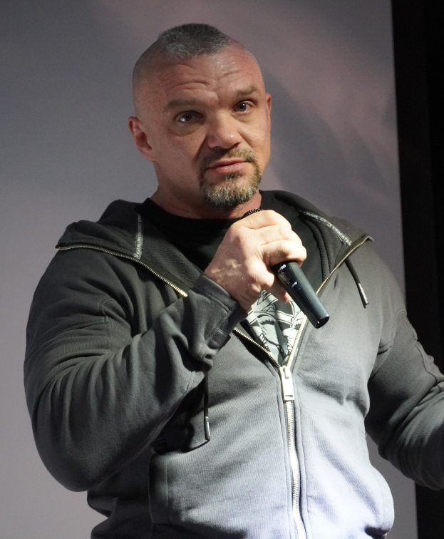 Ein Mann in einer schwarzen Kapuzenjacke und einem schwarzen Anzug mit einem weißen Hemd hält ein Mikrofon und steht selbstbewusst vor einer Wand, der Mann sieht aus wie ein Komiker mit zurückgekämmtem Haar.