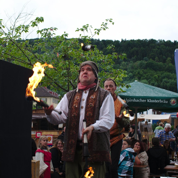 Eine Person mit Feuer steht in der Mitte einer Versammlung von Zelten und Ständen, mit Bäumen und Himmel im Hintergrund.