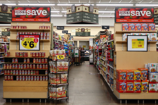Ein Innenraum eines Lebensmittelladens mit Regalen und Boden voller verschiedener Lebensmittel, Deckenbeleuchtung und einer nummerierten Anzeigetafel im Hintergrund, vor der eine Person steht.