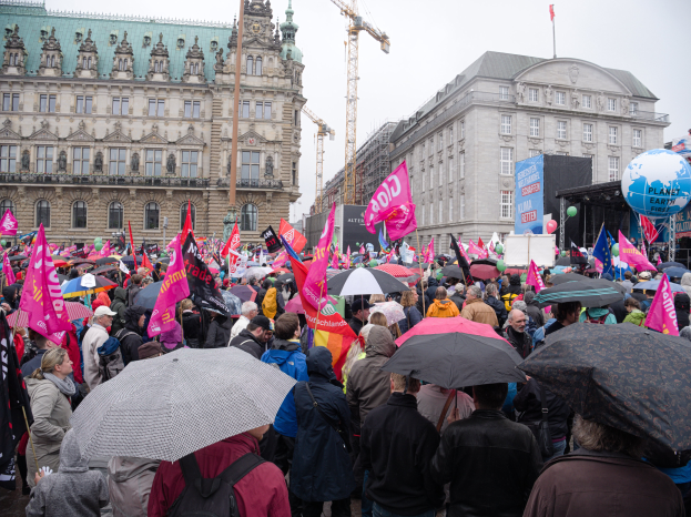 Eine große Gruppe von Menschen mit Schirmen und Transparenten nimmt an einer Women's March vor einem Gebäude in Berlin teil, mit Ballons und Stangen im Vordergrund und dem Himmel im Hintergrund.