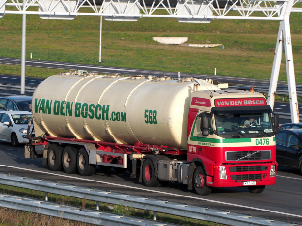 Ein großer weißer, roter und grüner Tanklastwagen fährt auf einer Autobahn, umgeben von anderen Fahrzeugen, mit einer Straßenbefestigung, Gras und einem Eisenrahmen im Hintergrund.
