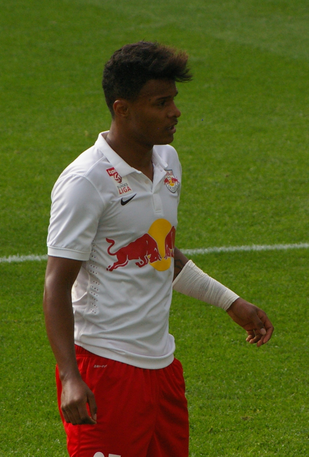 Ein Mann in weißem Hemd und roter Hose steht auf einem Fußballfeld, schaut nach rechts mit entschlossener Miene und sieht aus wie ein Flügelspieler.