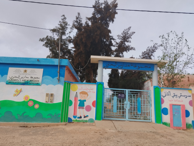 Blau-weißes Gebäude mit einer Wandmalerei, einem Tor, einer Namensschilder, Häusern, Bäumen, einem Mast mit Drähten und einem bewölkten Himmel, das als Eingang zu einer Kinder Schule dient.