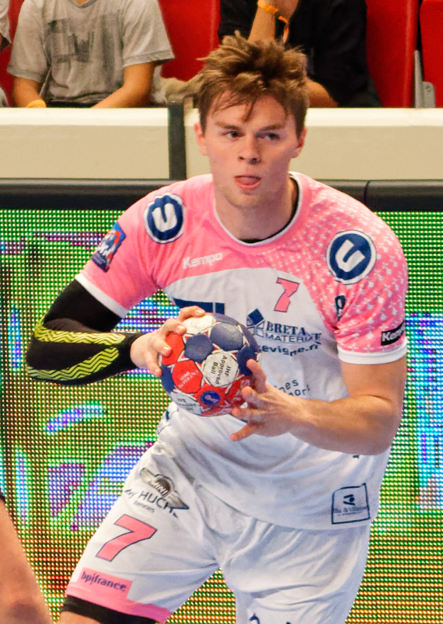 Ein Mann in einem pinken Hemd hält einen Fußball, mit der Hand eines anderen Menschen auf der linken Seite und ein paar Menschen im Hintergrund auf Stühlen sitzend.
