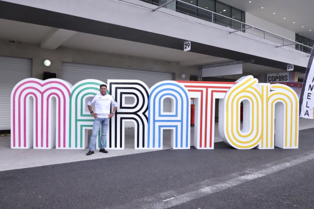 Ein Mann steht vor einem großen "Marathon"-Schild mit einem Banner rechts daneben, im Hintergrund ist ein Gebäude mit Lampen, Geländern und Fensterläden zu sehen.
