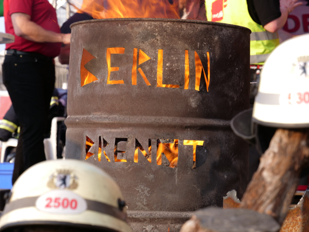 Eine Gruppe von Menschen steht um einen Feuerhydranten mit der Aufschrift "Berlin", mit einem brennenden Fass im Vordergrund, Helmen unten und einem hölzernen Objekt rechts.