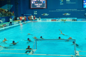 Eine Gruppe von Menschen spielt Wasserball in einem Pool mit einem Netz, umgeben von Zuschauern, Tafeln mit Text und Logos, Trägern und einem Bildschirm mit Zahlen und Text bei den FINA-Weltmeisterschaften 2015 in Kazan.