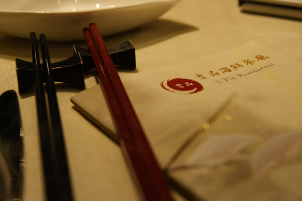 Ein Tisch mit einer Speisekarte, die "Ji Pin Restaurant" auf Mandarin und Englisch heißt, sowie zwei Paare Stäbchen (eines rot, eines schwarz) und eine weiße Schüssel vor den Stäbchen.