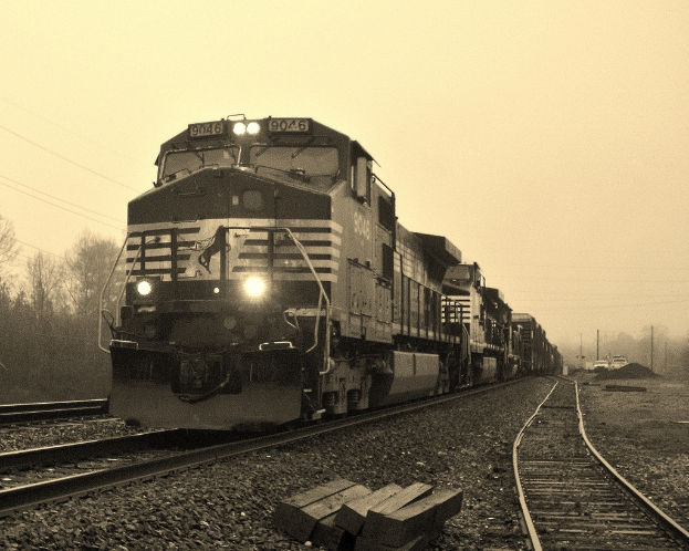 Ein Zug auf Eisenbahnschienen mit Autos, Strommasten, BÃ¤umen und einem Himmel im Hintergrund.