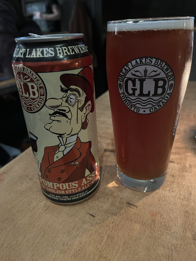 Eine hellgelbe Dose des Great Lakes Brewery-Pompous Ass-Biers und ein volles Glas goldener Bier auf einem hellen Holztisch, mit ein paar Leuten im Hintergrund.